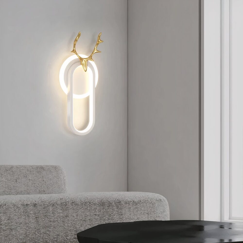Wandlamp met een lichtgevende witte cirkel en vooraan een lichtgevend wit ovaal met bovenaan een hertenkop in goud. Gemonteerd op een witte muur. Grijze bank en zwarte tafel.