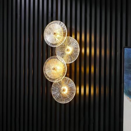 Wandlamp met gouden structuur die 4 lichtgevende glazen cirkels onder elkaar houdt. Gemonteerd op een zwarte muur met verticale ton sur ton strepen.