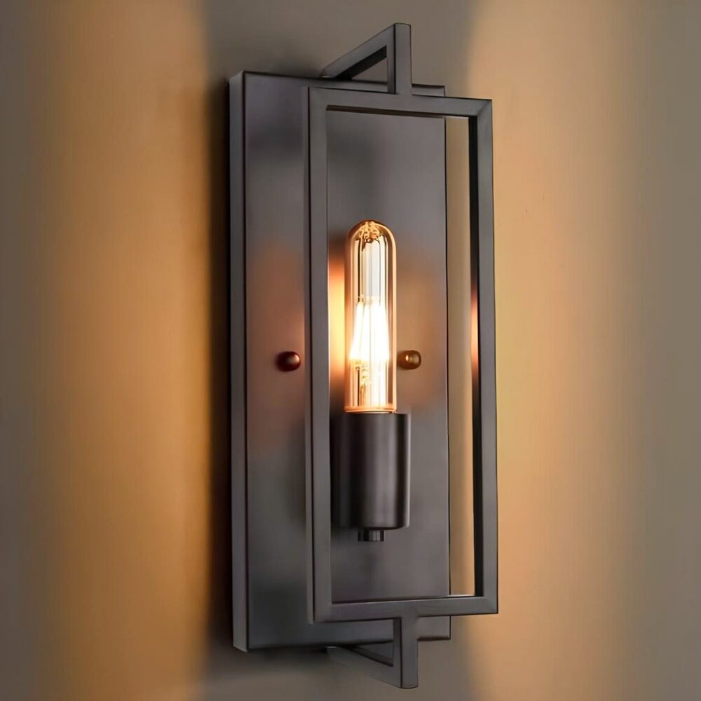 Wandlamp in de vorm van een rechthoekig industrieel frame van zwart metaal. De lange lamp brandt in het midden van het frame. De wandlamp is bevestigd aan een bruine muur.
