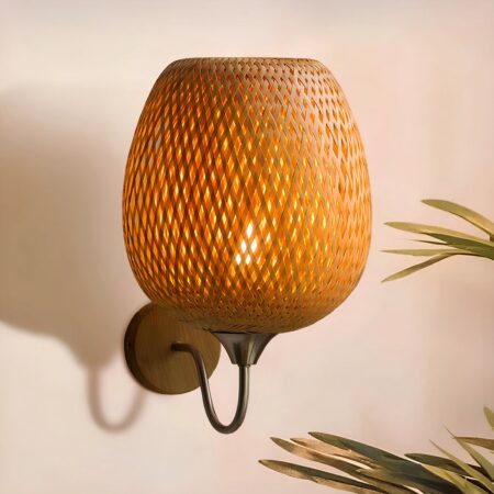 Rotan wandlamp met ronde houten steun bevestigd aan de muur en verbonden met de zwanenhals steel waar de ronde ovaalvormige rotan kap is bevestigd. De lamp brandt. Rechtsonder zijn enkele palmbladeren te zien.