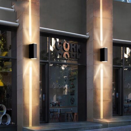 Winkelpui met etalages omlijst door betonnen pilaren waaraan een wandlamp is bevestigd. De wandlamp is rechthoekig en zwart van kleur en verspreidt een bundel warm licht boven en onder.