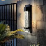 Buitenwandlamp in de vorm van een bubbelbuis in een zwarte aluminium structuur die is bevestigd aan een stenen muur. Links een groot zwart hek, een deel van een palmboom voor de muur en vallende regen.