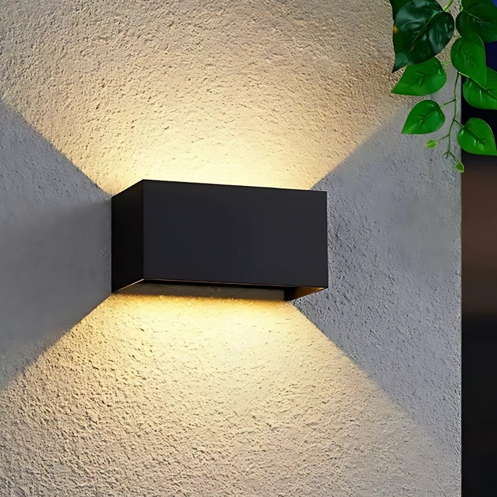 Zwarte rechthoekige buitenwandlamp met dubbele lichtbundel. Gemonteerd op een beige buitenmuur en ingeschakeld.